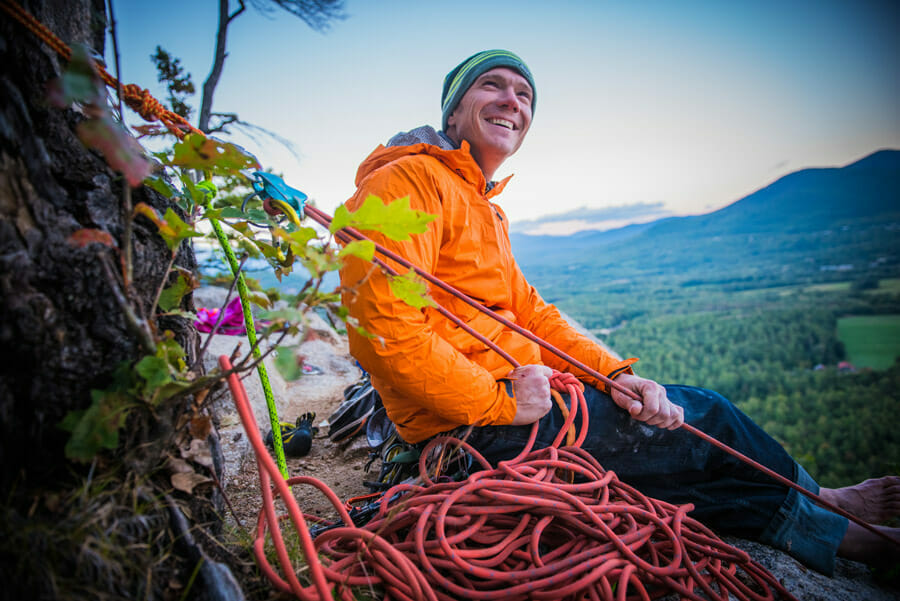 Person sitzt mit orangefarbener Softshelljacke auf Felsvorsprung beim Klettern.